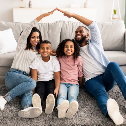 Happy family sitting together on a living room floor forming a roof shape with their hands, symbolizing homeowners insurance, home protection, and family security.